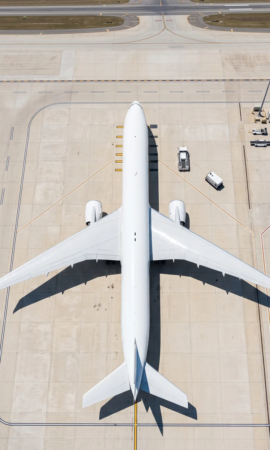 Overhead aerial view of a large unmarked white wide-body aircraft parked on a pale concrete airport apron, its fuselage running the length of the frame and wings spread symmetrically.