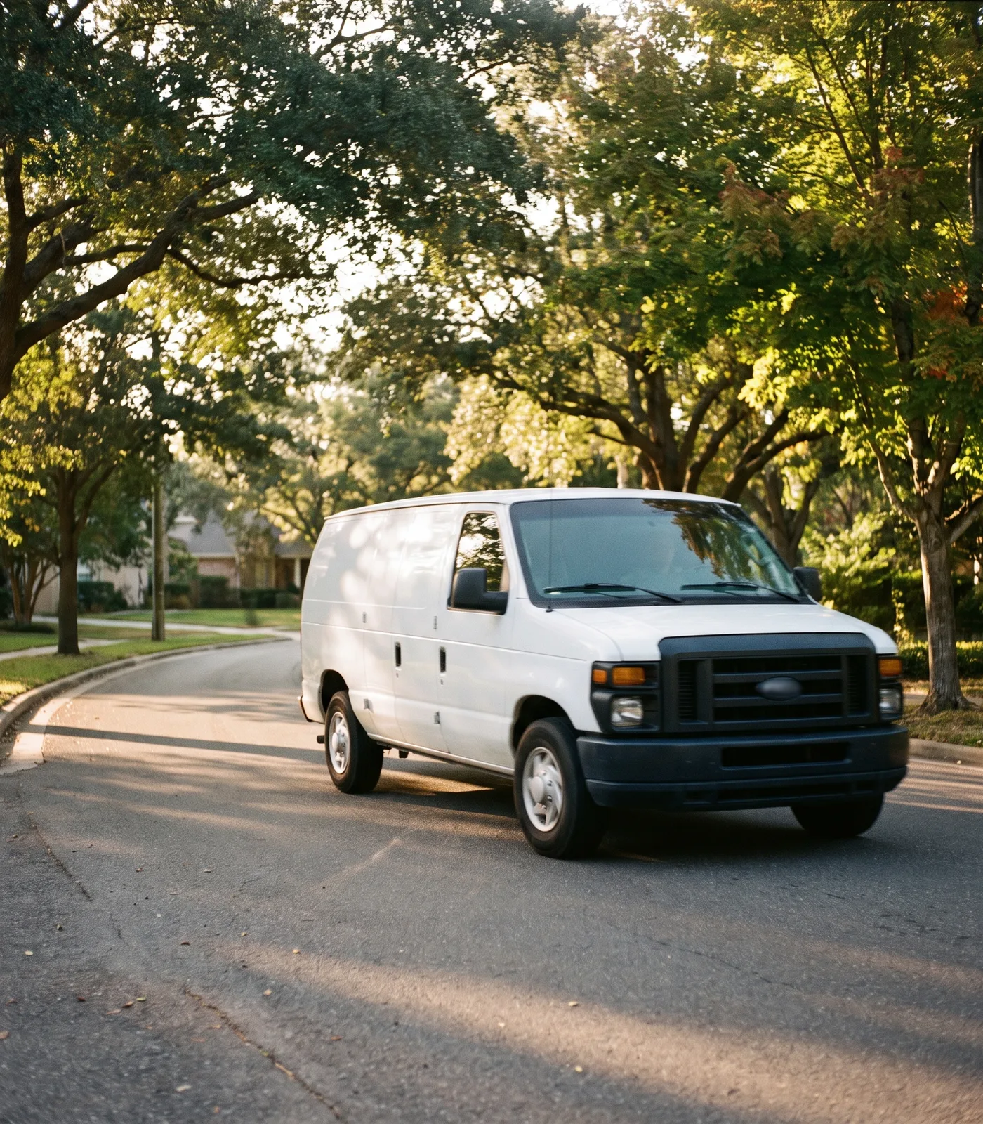 A warm daylight photograph of a plain white, unbranded service van driving along a quiet tree-lined suburban road.